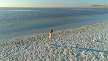 Young lonely woman in white dress is walking along shore of salt lake. Aerial view. video