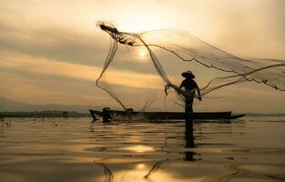 Silhouette of fisherman at sunrise, Standing aboard a rowing boat and casting a net to catch fish for food photo