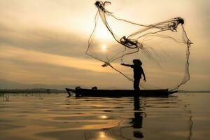 silueta de pescador a amanecer, en pie a bordo un remo barco y fundición un red a captura pescado para comida foto