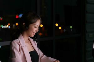 Young Asian woman broker international stock traders wearing headset working actively at night in office, Concept of customer support agent provide service on telephone. photo