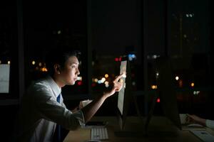 Young Asian broker international stock traders working at night in office using computer and thinking. photo