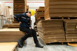 Warehouse worker sitting on the floor in front of stack of cardboard boxes with pain in the back due to frequently lifting heavy objects photo