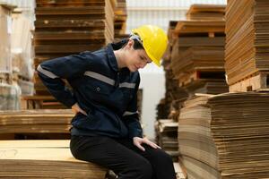 Warehouse worker sitting on the floor in front of stack of cardboard boxes with pain in the back due to frequently lifting heavy objects photo