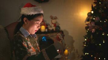 Asian woman sitting on the chair in front of the Christmas tree and opening a gift box, Merry Christmas and Happy Holidays photo