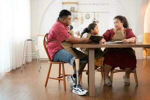 A plus-size family with a father wearing a prosthetic leg, is happily assisting a child with her homework and having fun together in the dining room of the house before cooking together photo