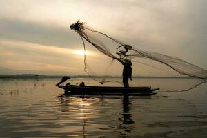 Silhouette of fisherman at sunrise, Standing aboard a rowing boat and casting a net to catch fish for food photo