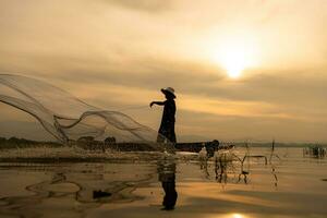 silueta de pescador a amanecer, en pie a bordo un remo barco y fundición un red a captura pescado para comida foto
