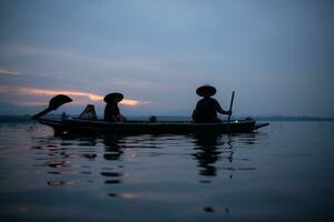 silueta de pescador a amanecer, en pie a bordo un remo barco y fundición un red a captura pescado para comida foto