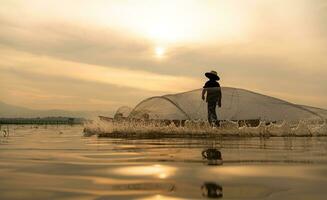 silueta de pescador a amanecer, en pie a bordo un remo barco y fundición un red a captura pescado para comida foto