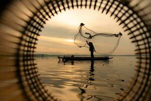 silueta de pescador a amanecer, en pie a bordo un remo barco y fundición un red a captura pescado para comida foto