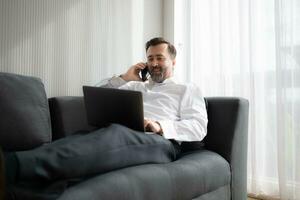 Businessman using laptop and talking on the phone while sitting on sofa at home photo