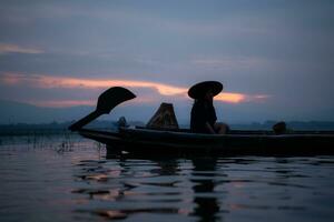 silueta de pescador a amanecer, en pie a bordo un remo barco y fundición un red a captura pescado para comida foto