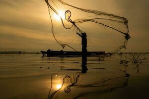 silueta de pescador a amanecer, en pie a bordo un remo barco y fundición un red a captura pescado para comida foto