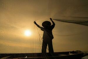 silueta de pescador a amanecer, en pie a bordo un remo barco y fundición un red a captura pescado para comida foto