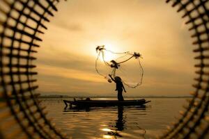 silueta de pescador a amanecer, en pie a bordo un remo barco y fundición un red a captura pescado para comida foto