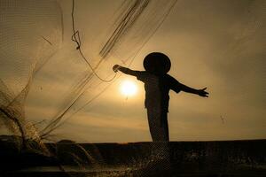 Silhouette of fisherman at sunrise, Standing aboard a rowing boat and casting a net to catch fish for food photo