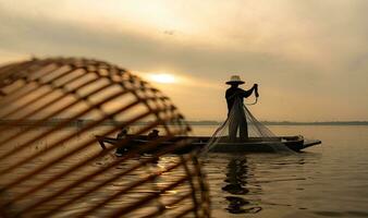 silueta de pescador a amanecer, en pie a bordo un remo barco y fundición un red a captura pescado para comida foto