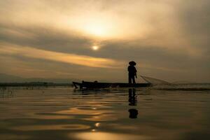 silueta de pescador a amanecer, en pie a bordo un remo barco y fundición un red a captura pescado para comida foto