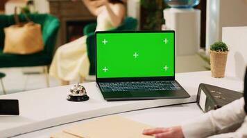 Employee checking greenscreen template on laptop, working at reception counter in lobby to help guests. Front desk staff looking at chromakey display with isolated blank template, seaside resort. photo