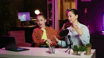 Cute kid teaching her mother how to do viral online dance moves while filming video for social media. Mom learning trending choreography from influencer daughter in apartment illuminated by pink neon