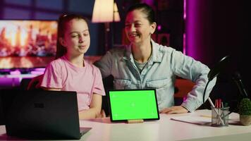 Cheerful child and loving mom record chroma key tablet review on online platforms. Young smiling daughter and parent presenting isolated screen device, filming in home studio with pink neon lighting video