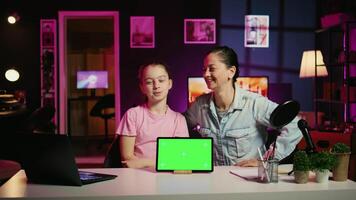 Smiling daughter filming online content with her parent using green screen tablet from sponsoring brand. Happy kid and her mother do internet videos for fans using chroma key device