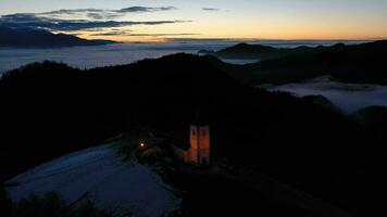Illuminated Church of St. Primoz and Felicijan in the Morning Twilight above the Clouds. Jamnik, Slovenia, Europe. Aerial View. Drone Flies Backwards and Upwards video