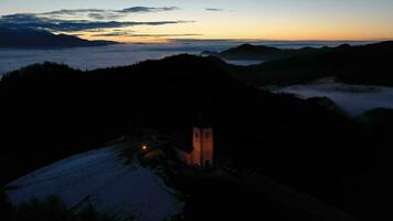 iluminado Igreja do st. primoz e Felicijan dentro a manhã crepúsculo acima a nuvens. jamnik, Eslovénia, Europa. aéreo visualizar. zangão moscas para trás e para cima video