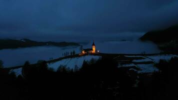 iluminado Iglesia de S t. primoz y felicijan a invierno noche brumoso crepúsculo. nublado cielo. jamnik, Eslovenia. aéreo vista. orbital video