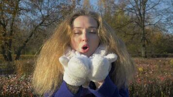 Young beautiful caucasian woman in white mittens is blowing out snow in sunny winter day in park. Slow motion. video