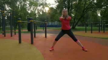 Jeune caucasien femme est élongation, est échauffement et est en gardant mains tout droit sur des sports sol dans ville parc à ensoleillé Matin. video