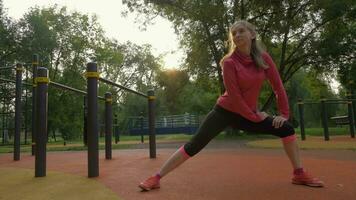 jovem caucasiano lindo mulher é alongamento e aquecimento em Esportes terra dentro verde parque às ensolarado manhã. lento movimento. video