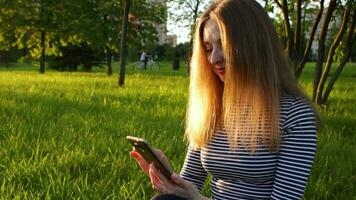 Young woman in striped shirt with long hairs is using smartphone and sitting on grass. Trees and cyclist in green city park at sunny evening on background. Mobile office concept.. video