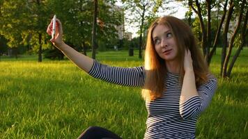 Jeune femme dans rayé chemise avec longue Cheveux est séance sur herbe et prise selfie par en utilisant téléphone intelligent. vert ville parc et des arbres à ensoleillé soir sur Contexte. video