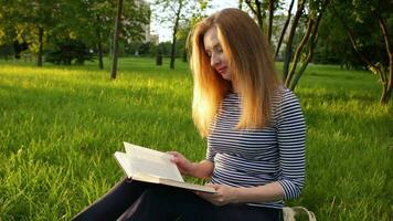 jovem mulher dentro listrado camisa com grandes cabelos é sentado em grama, lendo uma livro e girando uma página. verde cidade parque e árvores às ensolarado tarde em fundo. video