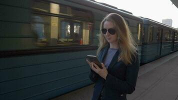 ung caucasian smal företag kvinna i blå jacka och solglasögon med lång hårstrån är vistas på metro station och använder sig av smartphone. tåg är avgår. video