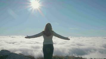 Woman with raised hands is standing on the edge of mountain above the clouds. video