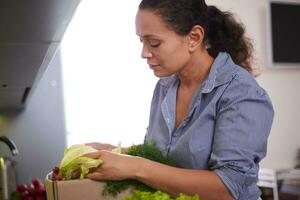 Close-up woman checking her online order list. Cardboard box with fresh vegetables and fruits standing by kitchen table photo