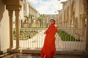 hermosa musulmán mujer con cabeza cubierto en un cabeza bufanda, a un mezquita, sonriente mirando a cámara, mediante su espalda foto