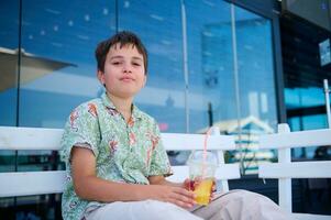 hermoso pre adolescente chico en verano camisa, participación un vaso de fresco exprimido naranja jugo, con confianza mirando a cámara foto