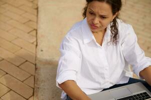 Mixed race young woman expressing sadness failing business tender project, looking aside, sitting with laptop outdoors photo
