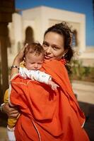 Beautiful Middle Eastern woman, loving mother in Oriental attire, holding her newborn baby boy, looking at camera photo