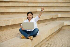 Happy female student of university remotely works on laptop, keeping arm raised, celebrates the successful pass of exams photo