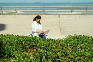 Businesswoman working on laptop outdoor, sitting on a bench against Atlantic ocean background photo