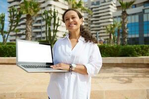 Smiling sales manager showing laptop mockup screen, standing against buildings background with developed infrastructure photo