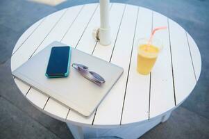 Top view. Laptop, mobile phone mockup screen, sunglasses and glass of fresh squeezed orange juice on white wooden table photo