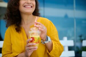 Details on hands of blurred pretty woman holding glass of freshly squeezed orange juice and cutely smiling looking aside photo