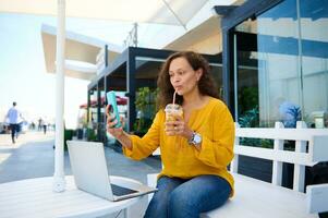 Beautiful woman blogger drinking orange juice and taking selfie, sharing her lifestyle on social media content. Blogging photo