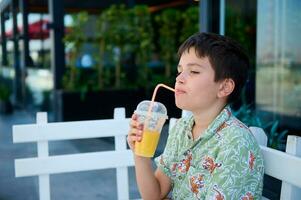 Side portrait of an adorable handsome Caucasian teen boy in summer clothes drinking freshly squeezed juice from a straw photo