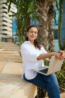 Young adult woman in casual clothes, sitting on a stone bench with laptop on her knees, smiling cutely looking at camera photo